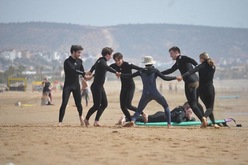 A group of people in wetsuits on a beach, holding hands in a circle. 3