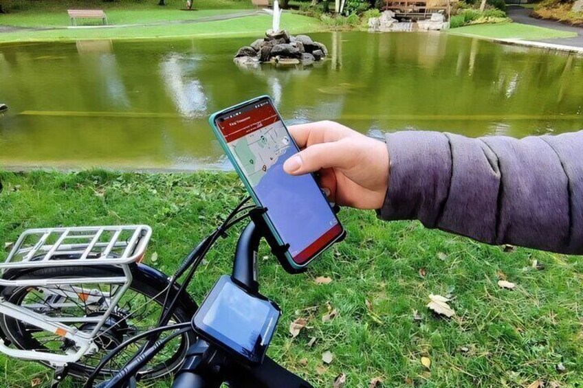 Iemand houdt een smartphone vast met een kaart-app, een fiets met een mandje, en een vijver met een fontein op de achtergrond. 2