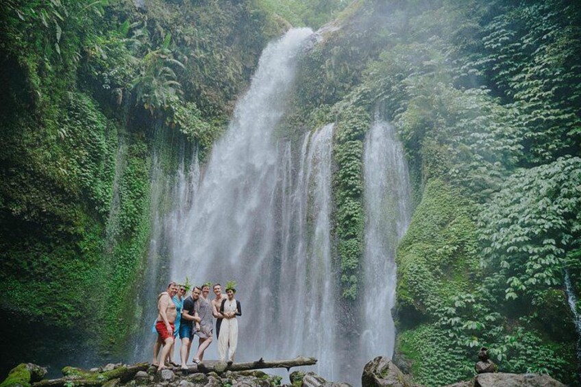 A waterfall with lush greenery and a group of people standing on a log. 1
