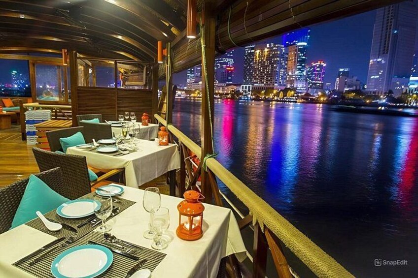 A dining area with tables set for a meal, overlooking a river with a city skyline in the background. 3