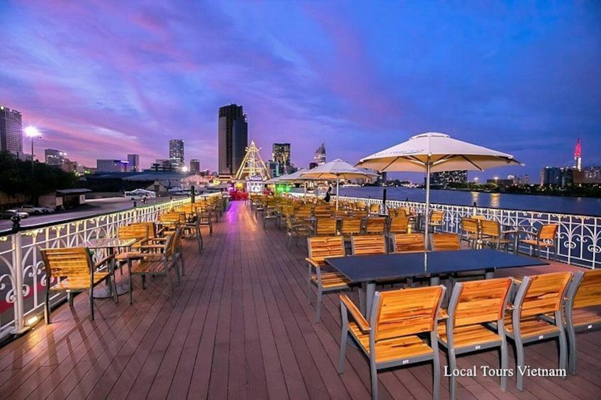 A waterfront deck with wooden chairs and tables, a Ferris wheel, and city skyline in the background. 1