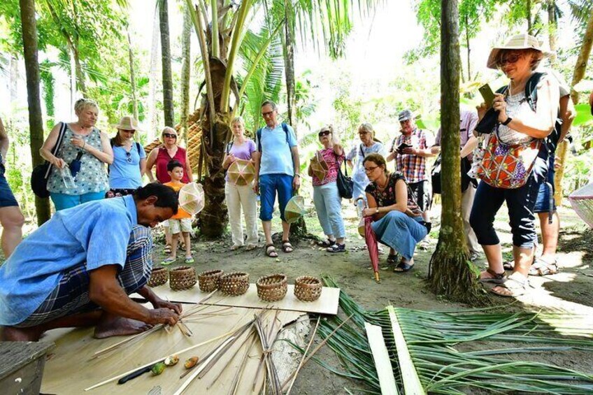 A group of people observing a person weaving baskets in a tropical setting. 1