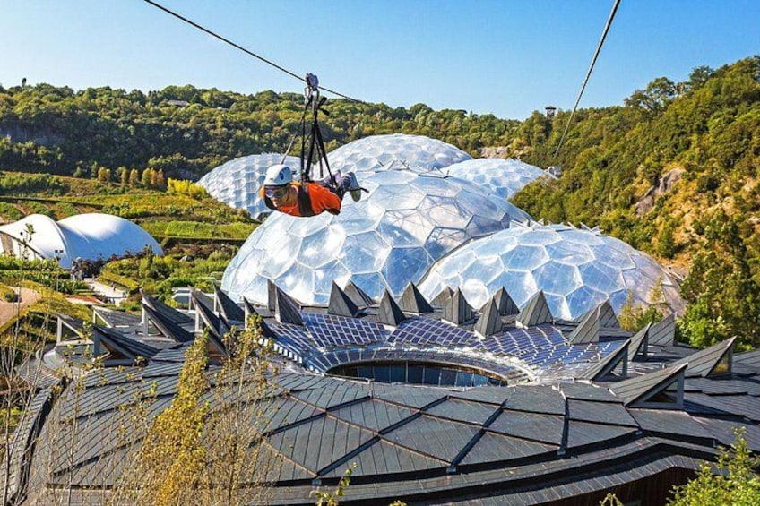 Een persoon hangt aan een kabel boven een gebouw met een koepelvormig dak, omringd door zonnepanelen en een bergachtig landschap. 4