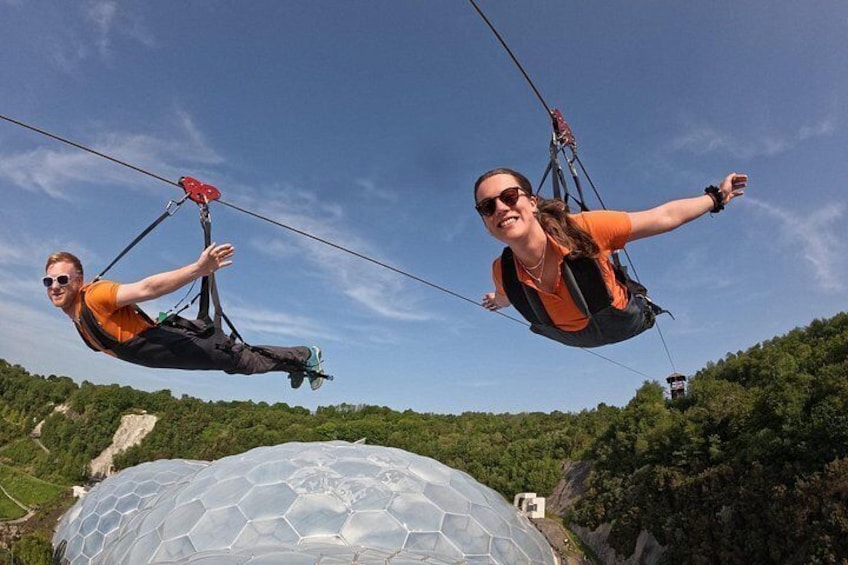 Two people suspended in the air by a cable, with a large dome structure and a forested hill in the background. 3