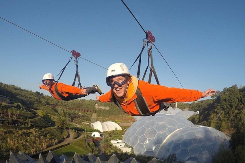 Two people suspended in mid-air by cables, wearing safety helmets and harnesses, with a large dome structure and a landscape with trees and a winding road below. 1
