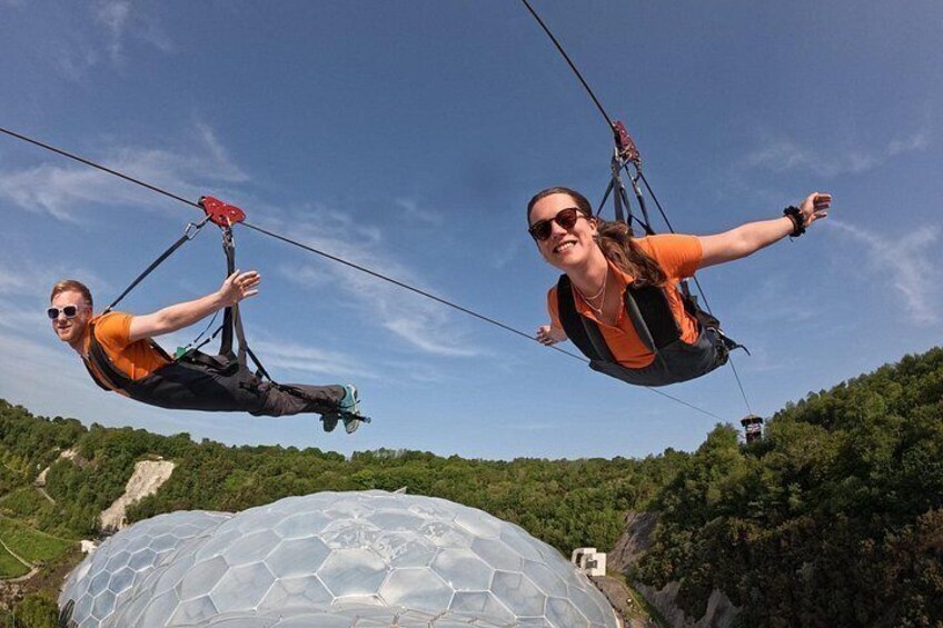 Two people suspended in the air by a cable, with a large dome structure and a forested hill in the background. 3