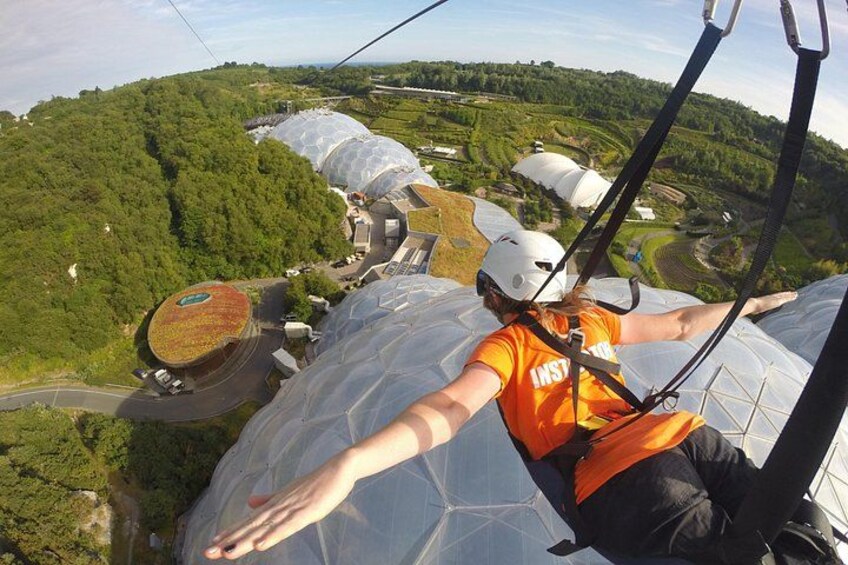 Een persoon in een oranje shirt en helm, met een tuig gespannen, kijkt uit over een landschap met groene heuvels en grote, koepelvormige constructies. 3