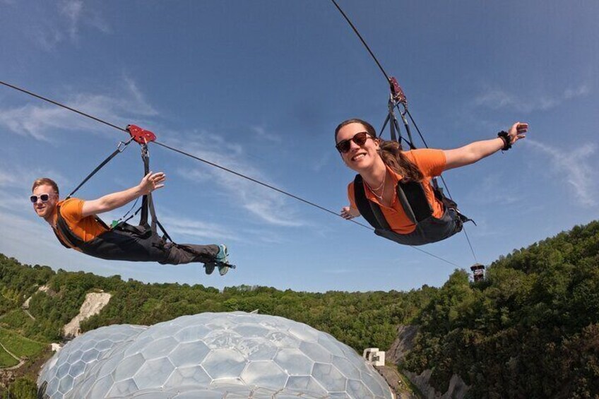 Two people suspended in the air by a cable, with a large dome structure and a forested hill in the background. 3
