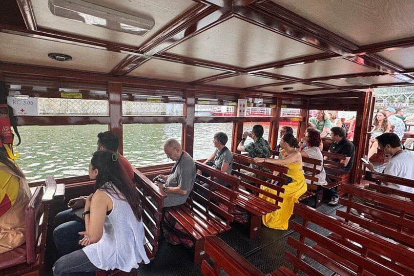 A boat interior with wooden benches and tables, passengers seated and standing, and a view of the water outside. 5