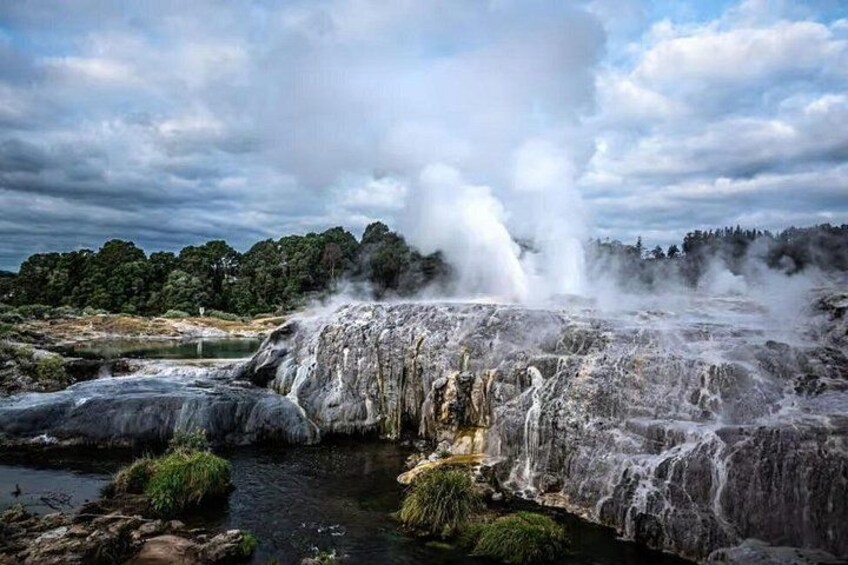 A geyser with steam erupting, surrounded by rocky terrain and a forested area. 3