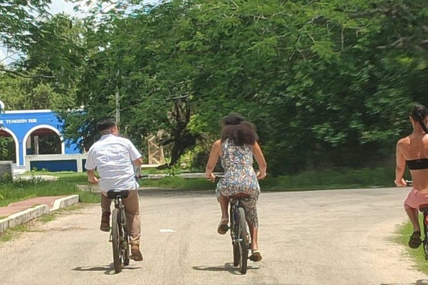 Three people riding bicycles on a paved road with trees and a blue building in the background. 5