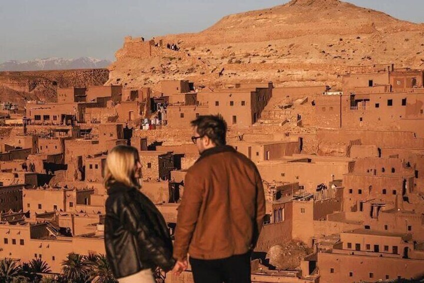 Un couple se tient devant un village traditionnel construit en briques de terre, avec une montagne en arrière-plan. 1