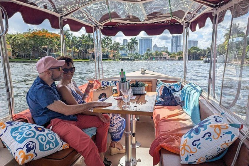 A couple on a boat with a table, cushions, and a view of the water and buildings. 5