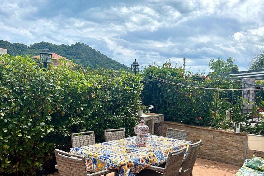 A patio with a table covered in a colorful tablecloth, surrounded by wicker chairs, and a lush green hedge in the background. 4