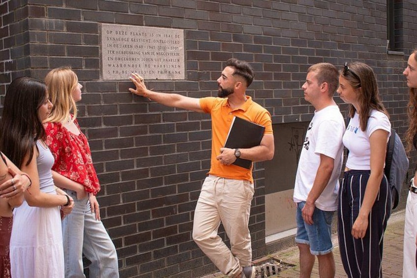 A man in an orange shirt gesturing towards a brick wall with a sign. 1
