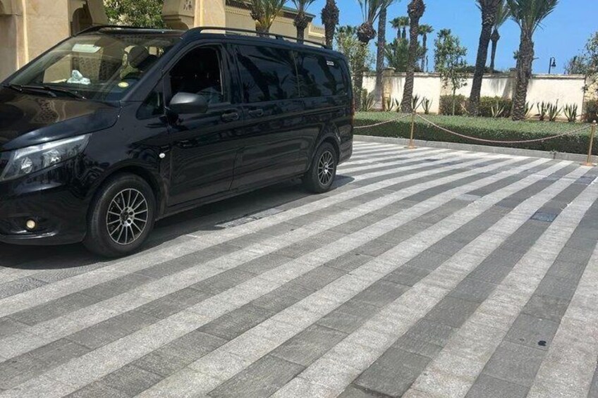 Een zwarte minibus geparkeerd op een gestreepte oprit, met palmbomen en een gebouw op de achtergrond. 5
