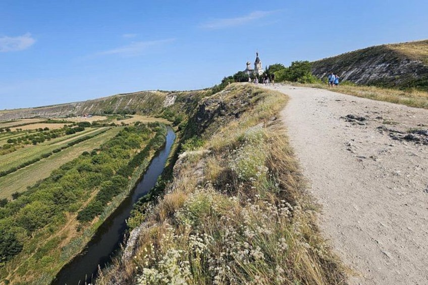 A dirt path alongside a river, with a small building and people in the distance. 1