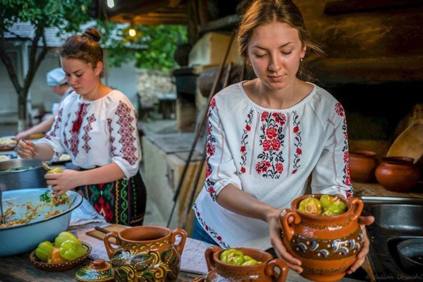 Two people preparing food outdoors, wearing traditional embroidered clothing, with cooking utensils and dishes visible. 2