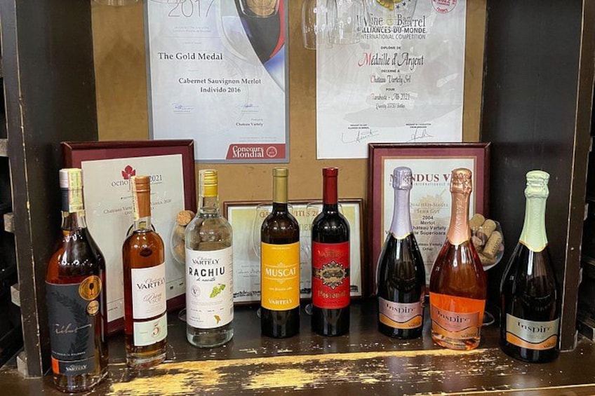 A selection of wine bottles on a wooden counter, with awards and certificates in the background. 2
