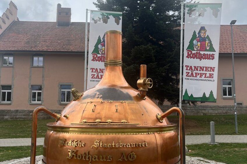A large copper kettle with inscriptions, set against a building with banners. 1