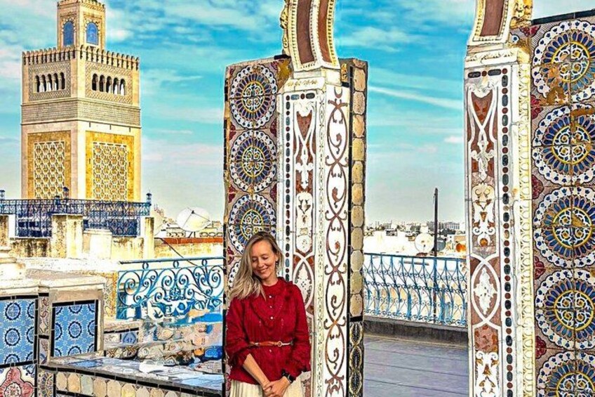 A person standing in front of a decorative gate with intricate patterns, a tower in the background, and a blue railing. 1
