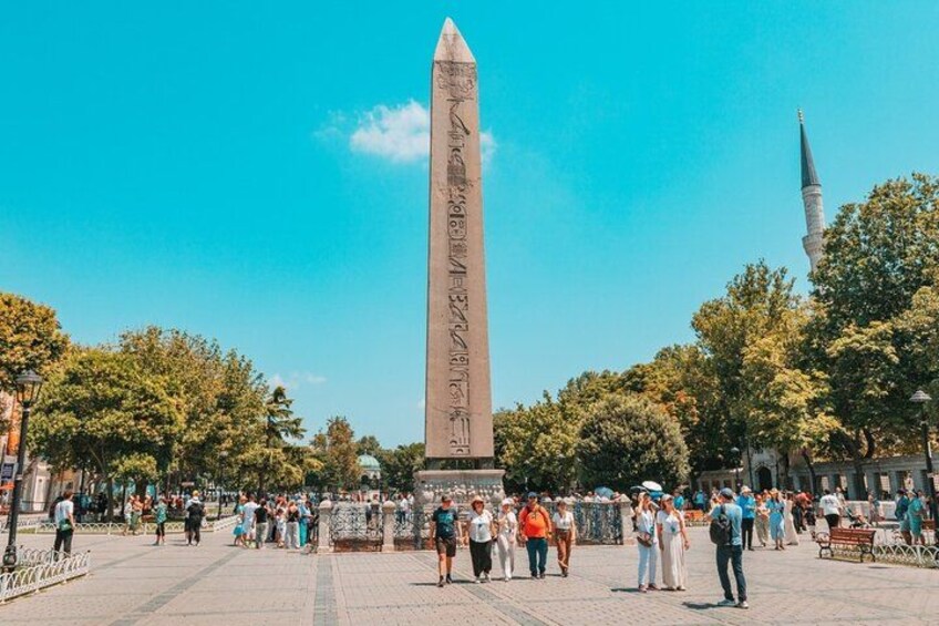 A tall obelisk with hieroglyphs, surrounded by trees and people in a public square. 3