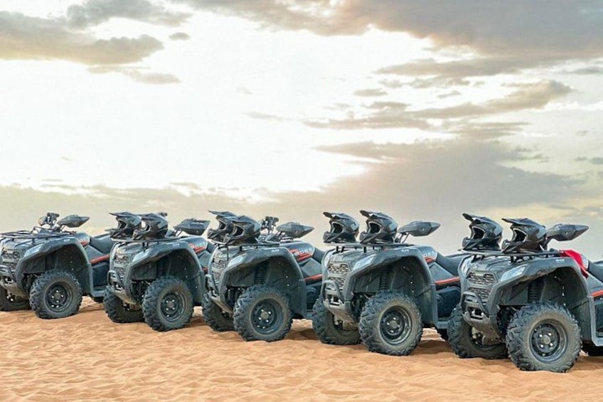 A row of ATVs parked on a sandy surface with a cloudy sky in the background. 2