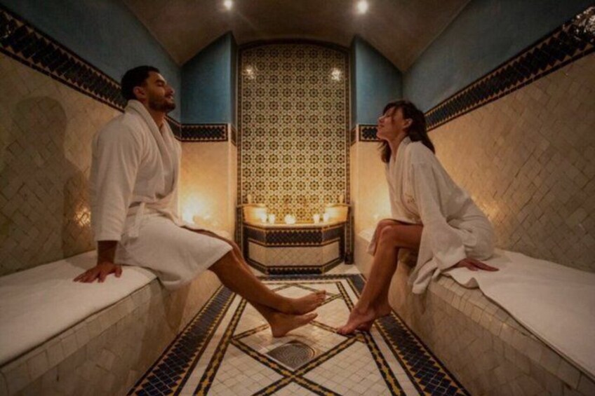 Two people in white robes sitting on benches in a room with patterned tile walls and a tiled floor. 1