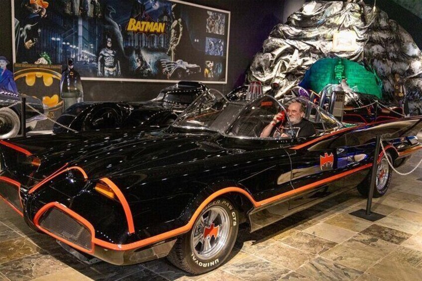 Ein schwarzes Auto mit orangefarbenen Akzenten und einem Batman-Logo, ausgestellt in einem Museum. Im Hintergrund ist ein Batman-Poster zu sehen. 5