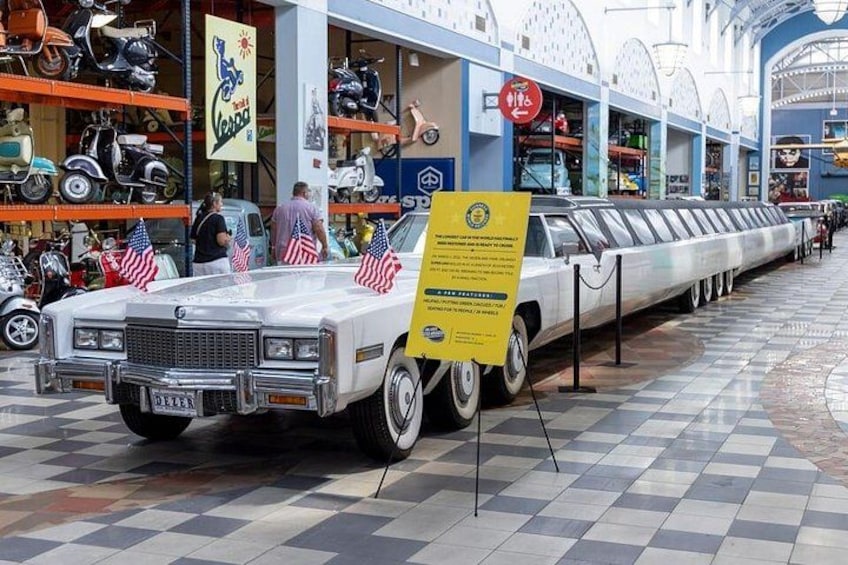 Ein Oldtimer wird in einer Ausstellung präsentiert, umgeben von verschiedenen Rollern und Motorrädern, mit amerikanischen Flaggen geschmückt. 2
