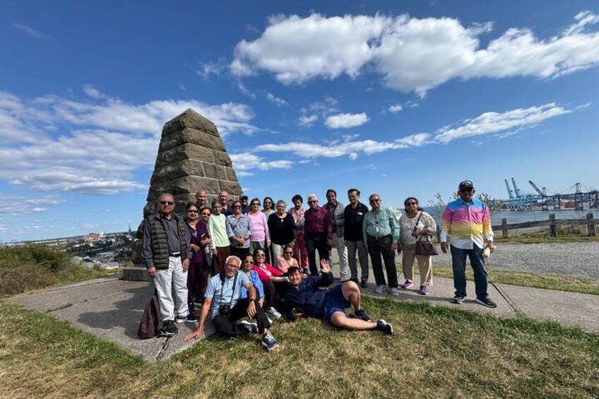 A group of people standing and sitting around a stone monument near a waterfront with a bridge in the background. 3