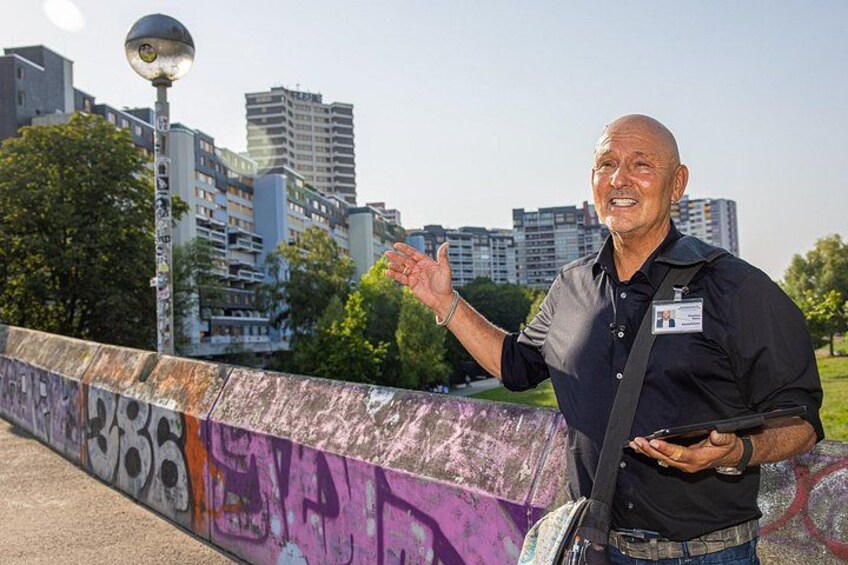 En man står på en bro med graffiti på väggen, en stadsbild syns i bakgrunden och himlen är klar. 1