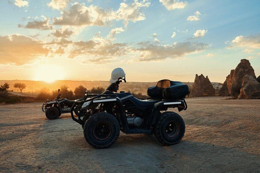 An off-road vehicle with a rider in a white helmet, a backpack, and a helmet on the back, parked on a dirt road with a sunset and rock formations in the background. 2