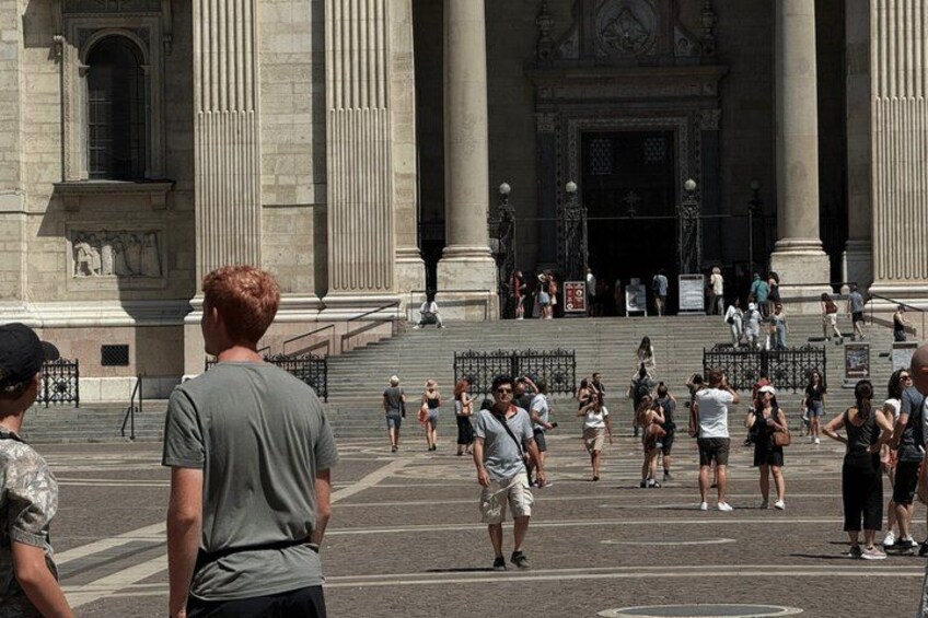 A large open plaza with people walking and standing, a grand building with columns in the background, and a clear sky. 4