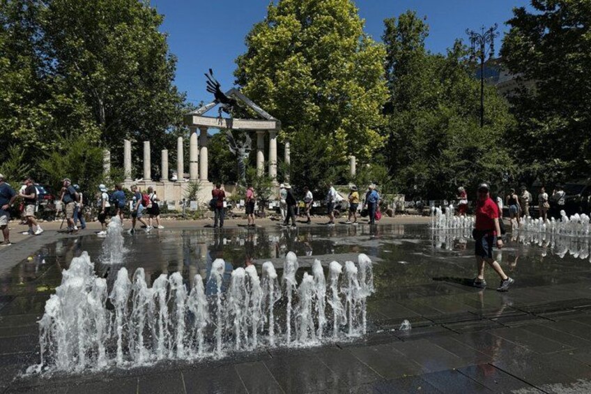 A public fountain with water jets, surrounded by a park with trees and a classical monument. 2