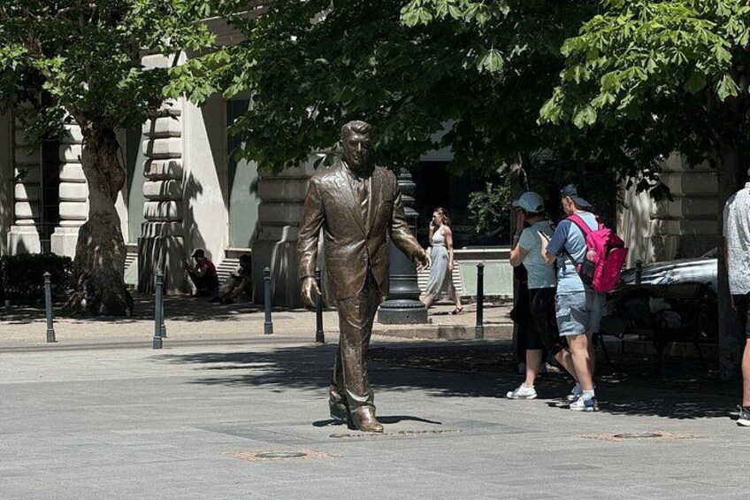 A bronze statue of a man in a suit, a tree, and people walking in a public square. 2