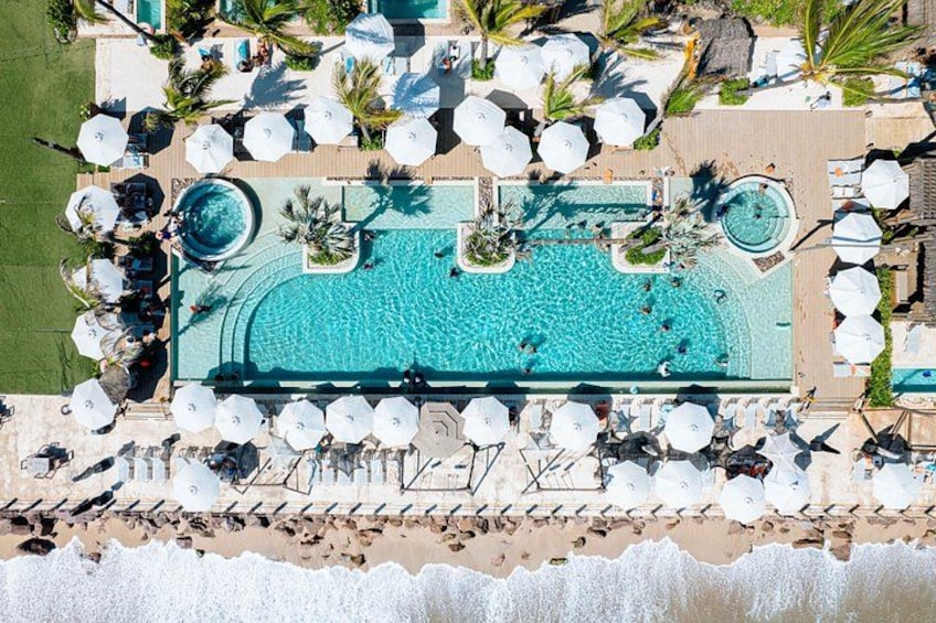 An aerial view of a resort with a large swimming pool surrounded by lounge chairs and umbrellas, with a beach in the foreground. 2