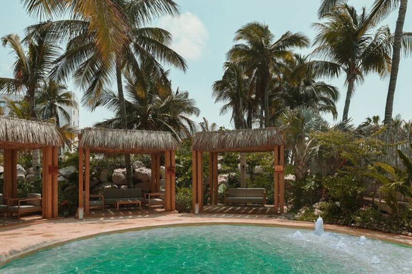 A pool with a water fountain, surrounded by palm trees and wooden gazebos. 5
