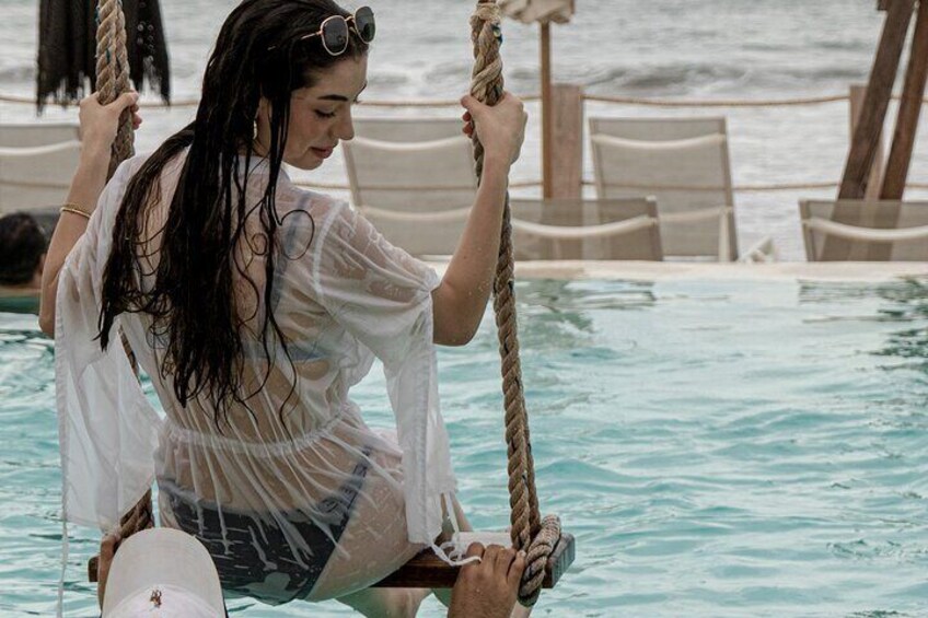 A person sitting on a rope swing over a pool with a beach in the background. 2