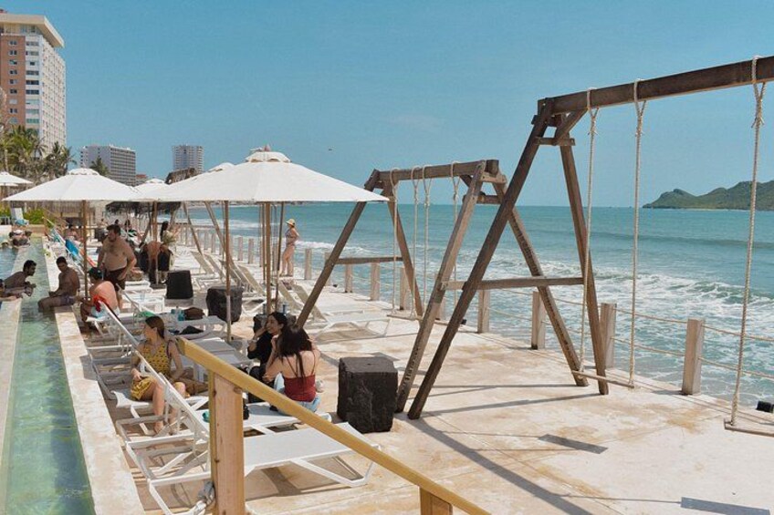 Ein Poolbereich am Strand mit Liegestühlen und Sonnenschirmen, einer Holzschaukel und Blick auf den Ozean. 2