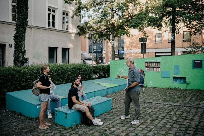 Eine Gruppe von Menschen sitzt auf blauen Bänken in einer Außenanlage mit einer grünen Wand und einem Gebäude im Hintergrund. 3