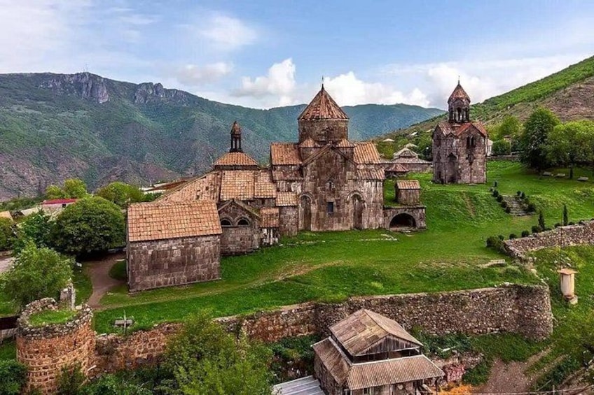 A historic church complex with multiple buildings, including a central church with a prominent dome and smaller structures, set against a mountainous backdrop. 3