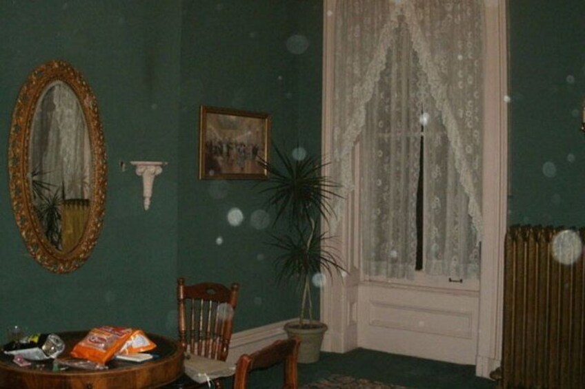 Una habitación con paredes verdes, una silla de madera, una mesa con objetos, un espejo, una ventana con cortinas de encaje y un radiador. 2