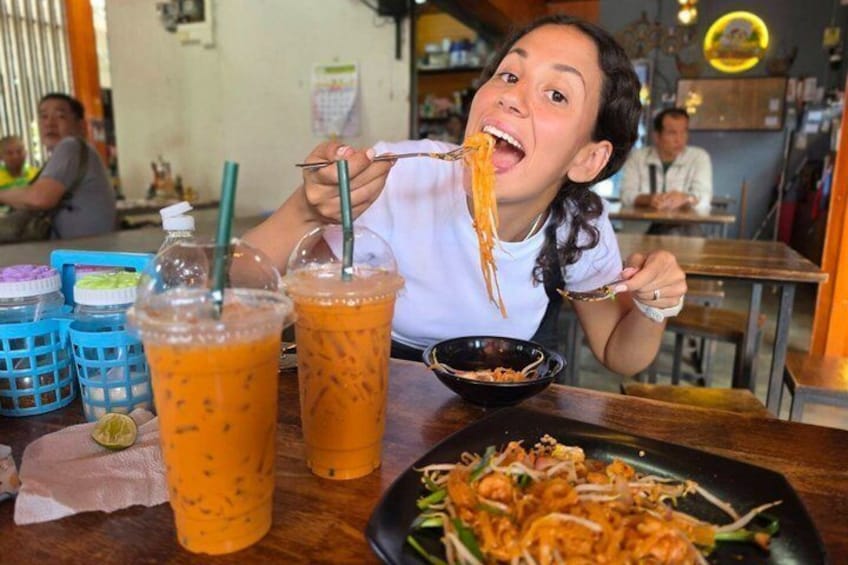 A person eating noodles with a side of soup and two iced drinks on a table. 1