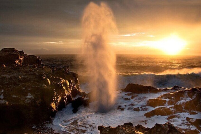 Un géiser en erupción al atardecer, con terreno rocoso y olas del océano. 3