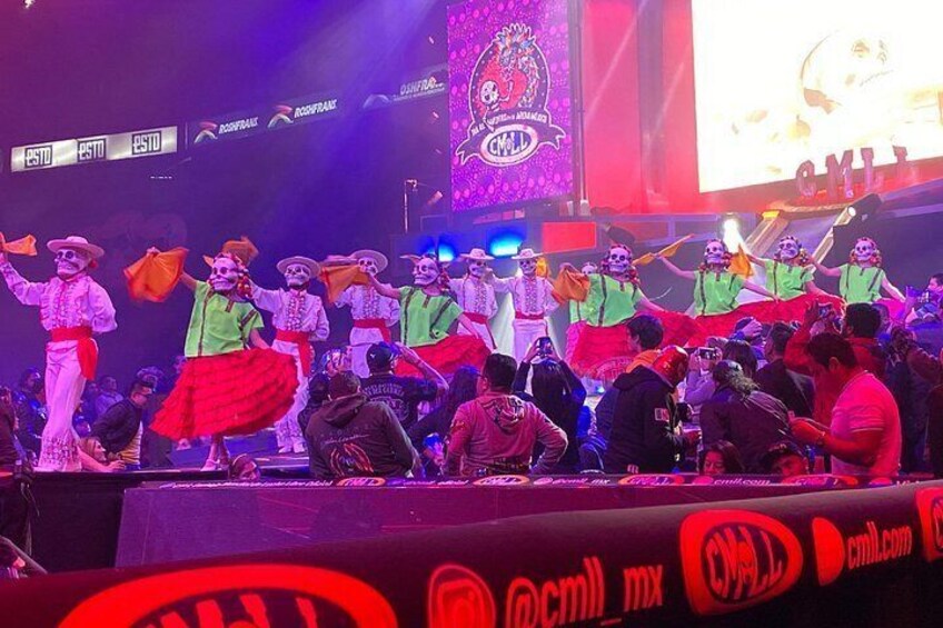 Un escenario con artistas vestidos con trajes tradicionales mexicanos, una pantalla grande mostrando texto y una multitud de espectadores. 4