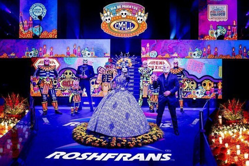 Un escenario con artistas vestidos con atuendo tradicional mexicano, un arreglo floral grande y el texto 'ROSHFRANS' en el suelo. 1