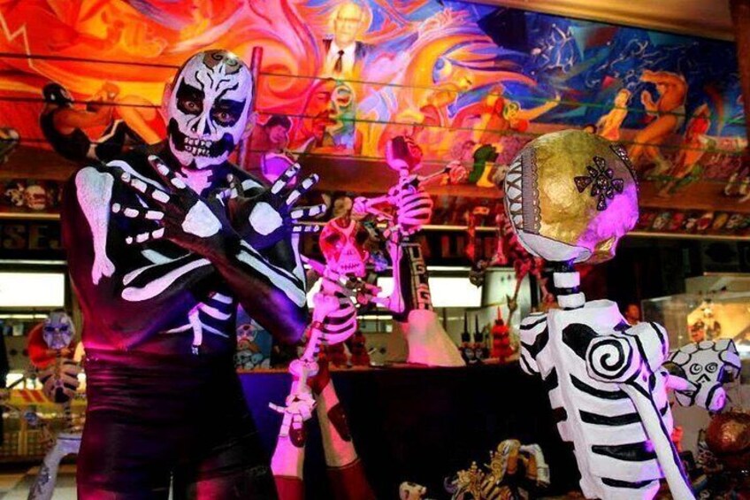 Una persona disfrazada de esqueleto sostiene un objeto dorado grande, con un mural colorido de fondo. 2