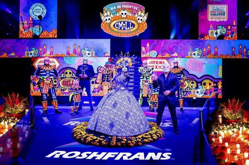 Un escenario con artistas vestidos con atuendo tradicional mexicano, un gran emblema circular, y el texto 'DIA DE MEXICO' y 'ROSHRANS'. 1
