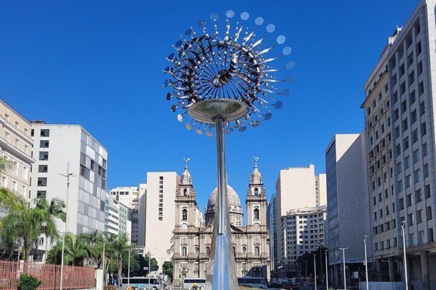A cityscape with a prominent sculpture in the foreground and a historic building with domes in the background. 2